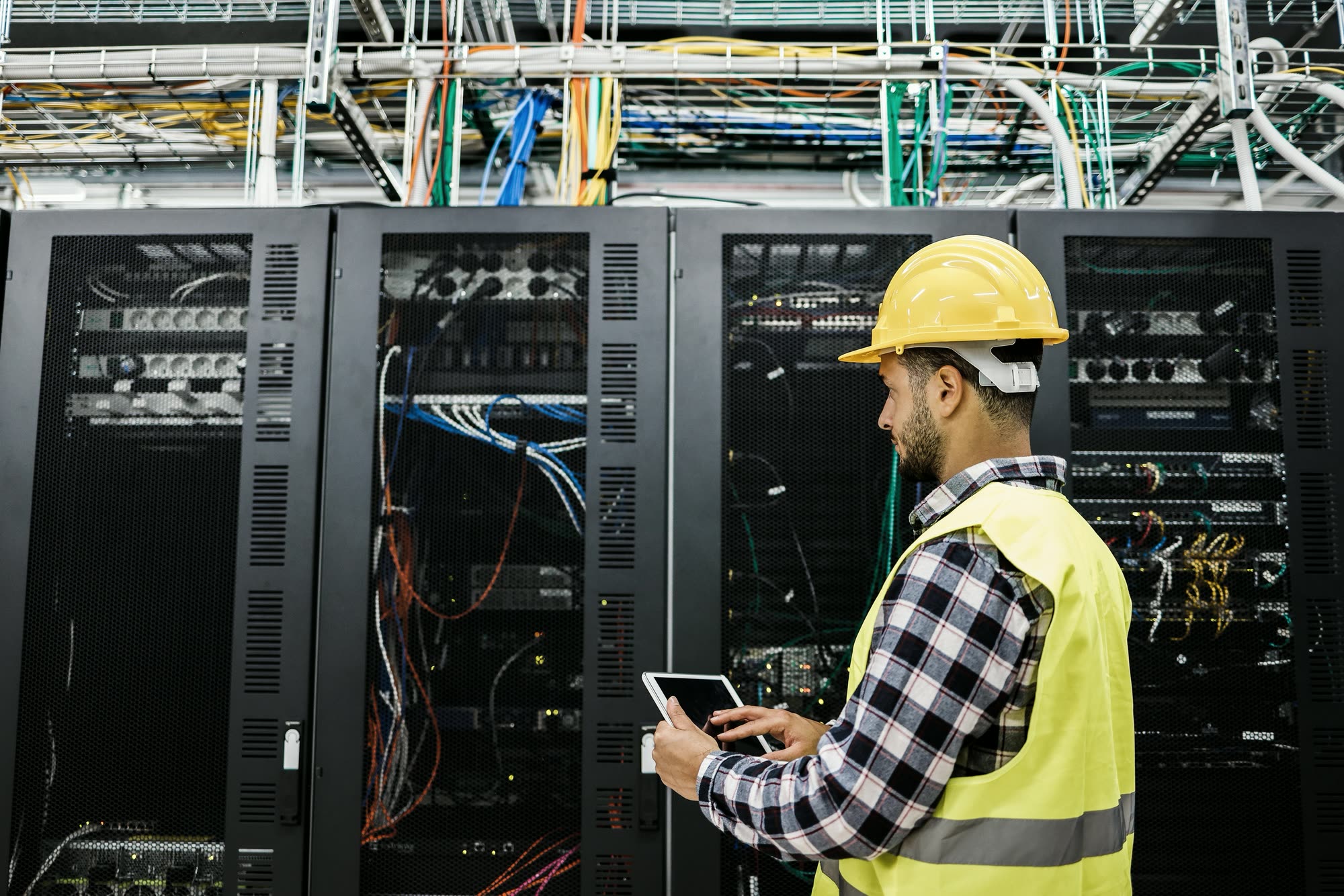 An engineer in a hard hat checking a server rack on a tablet
